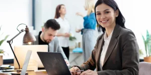 Professional woman smiling at laptop in a busy office environment — how to secure a job