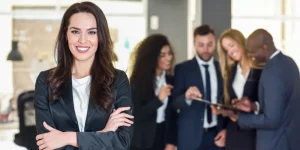 Confident professional woman standing with arms crossed and colleagues in the background - why you should keep a work journal for career growth